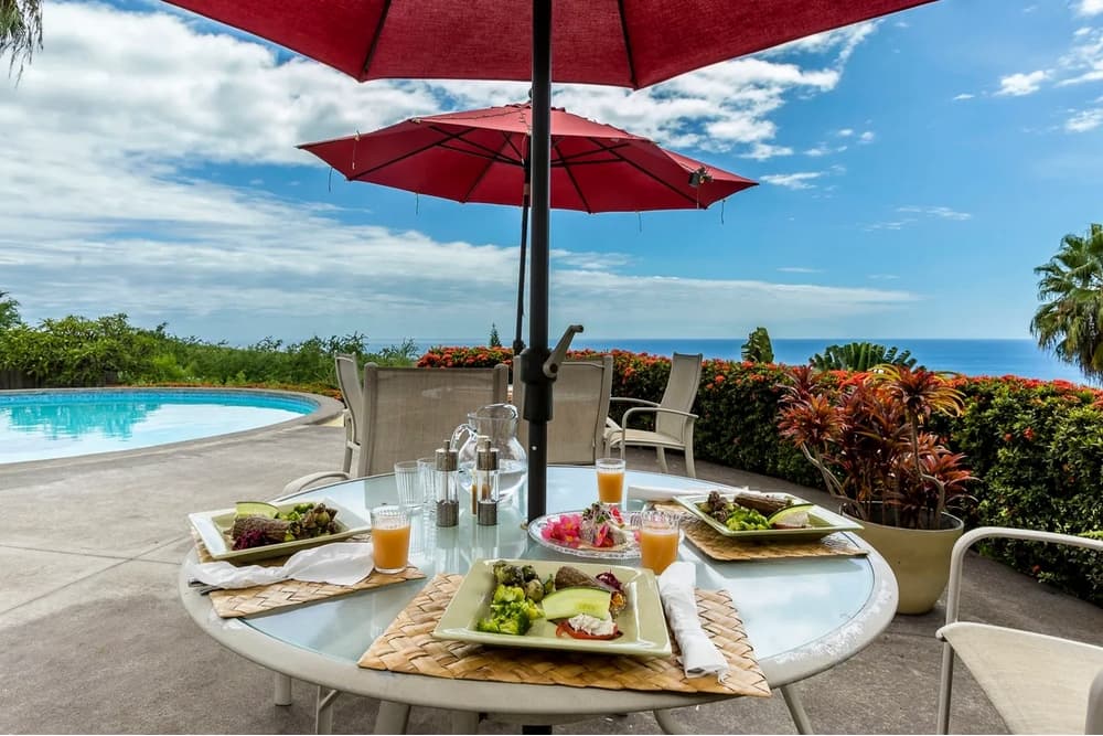 A table set with colorful meals and drinks under a red umbrella near a swimming pool and ocean view.