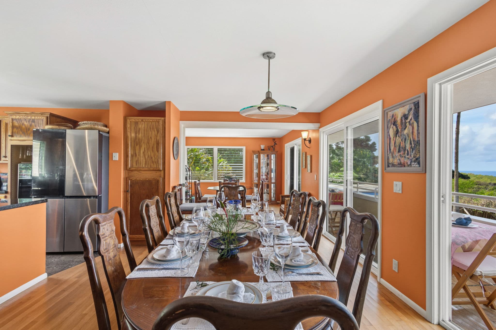 A spacious dining area featuring a long wooden table set for a meal, with orange walls and large windows offering a view.