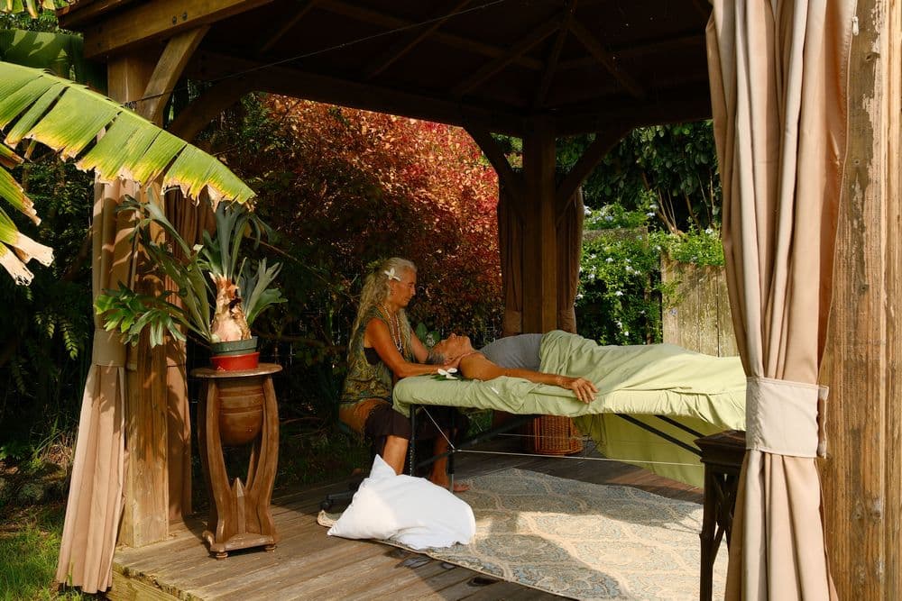 A woman provides a massage outdoors under a wooden gazebo surrounded by greenery.