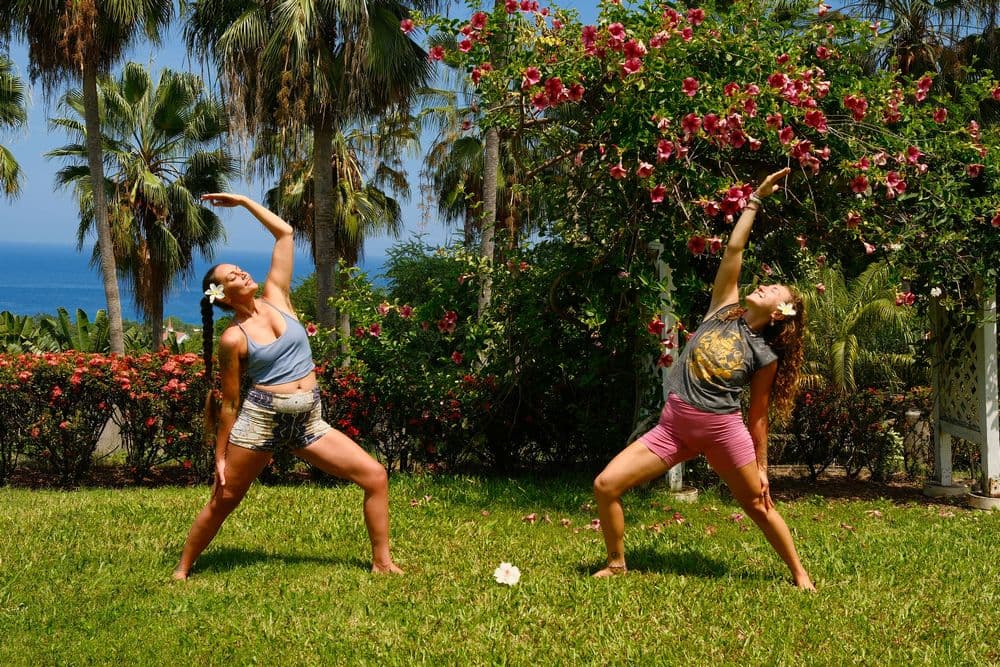 Two women practice yoga poses in a lush garden with blooming flowers and a view of the ocean.
