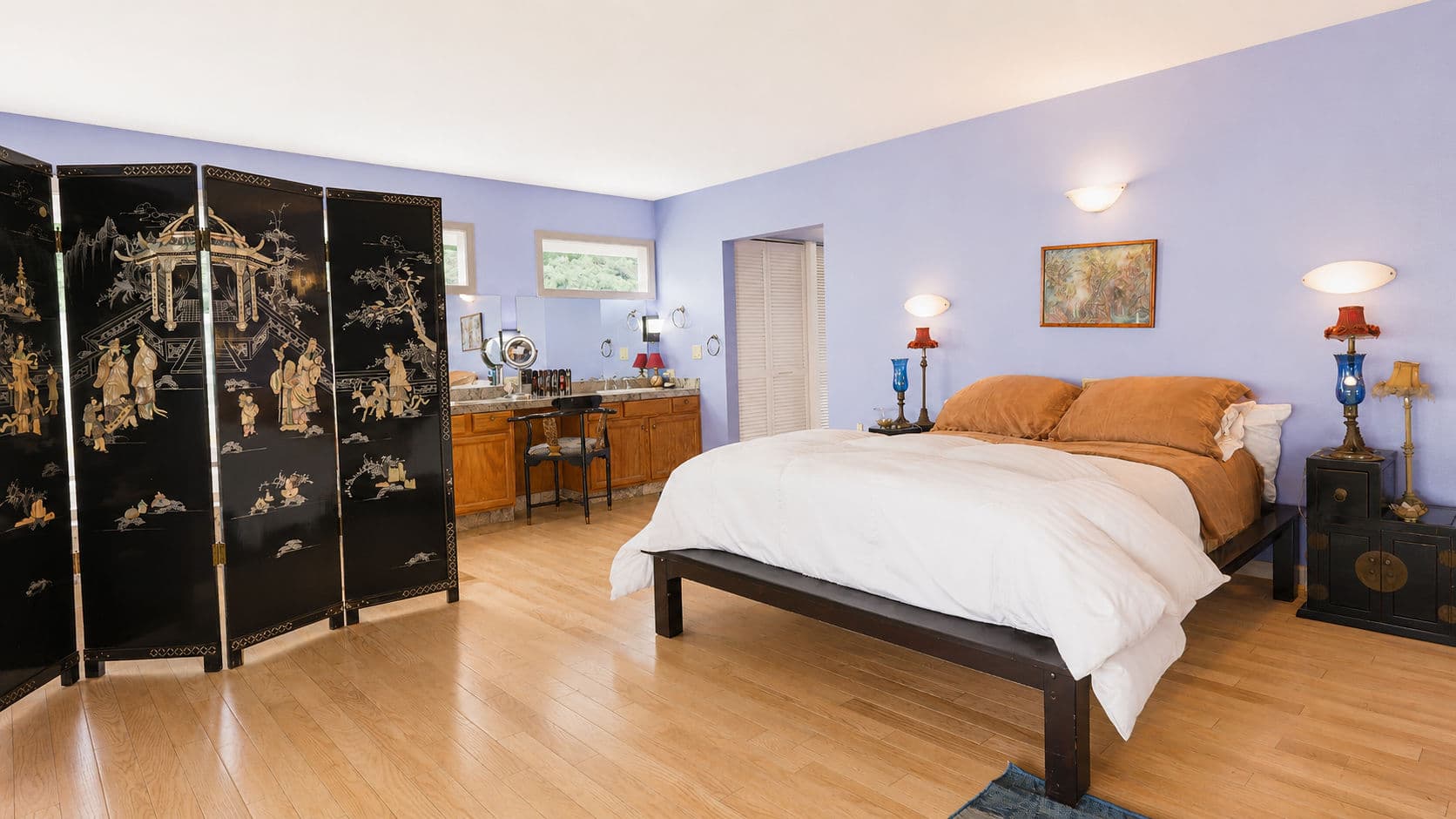 A spacious, serene bedroom featuring a bed with white bedding, a decorative black folding screen, and blue walls.