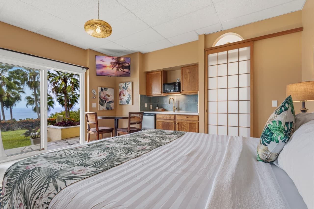 Cozy bedroom with tropical decor, kitchenette, and sliding doors opening to a view of palm trees.