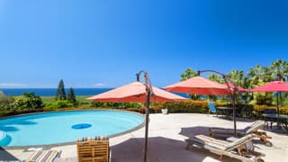 A scenic poolside area with lounge chairs and umbrellas overlooking a lush landscape and ocean under a clear blue sky.