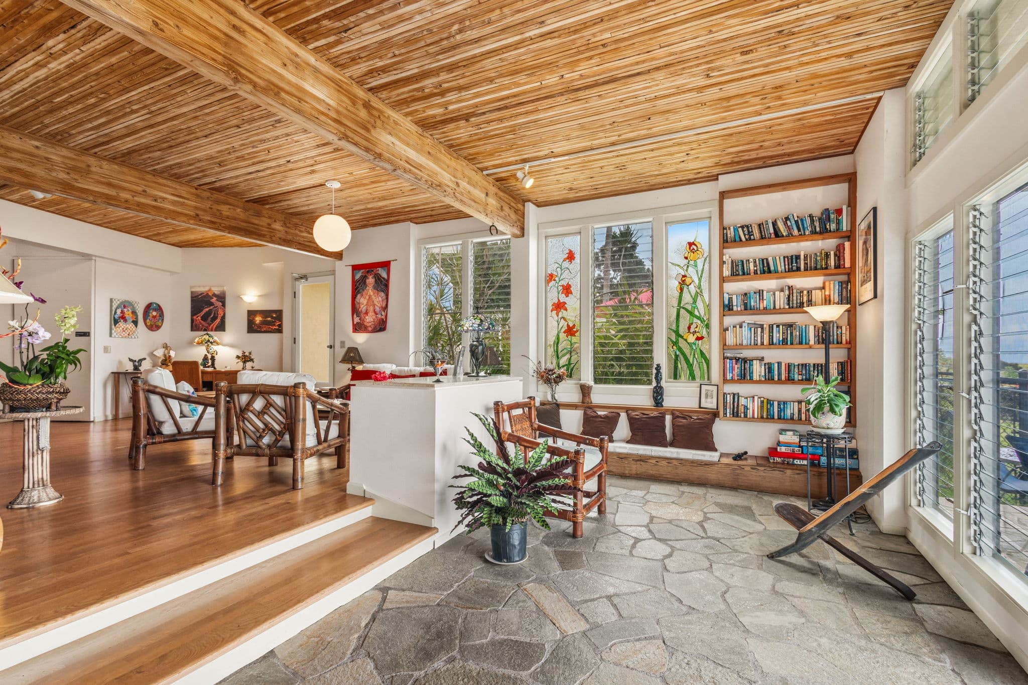 A bright, open living space featuring wooden beams, a stone floor, bookshelves, and large windows with greenery.