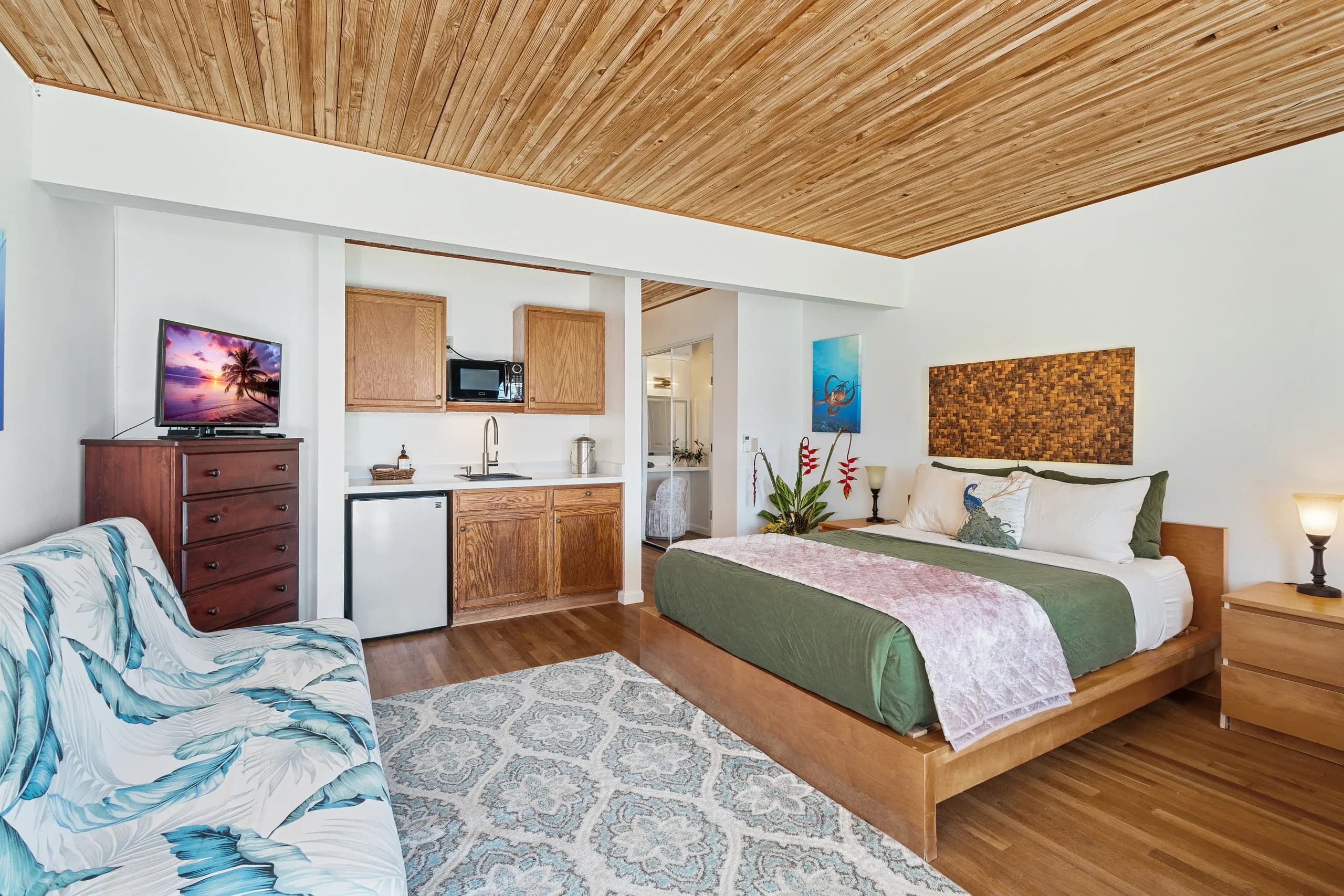 A cozy hotel room featuring a bed, kitchenette, and a sitting area with tropical decor.