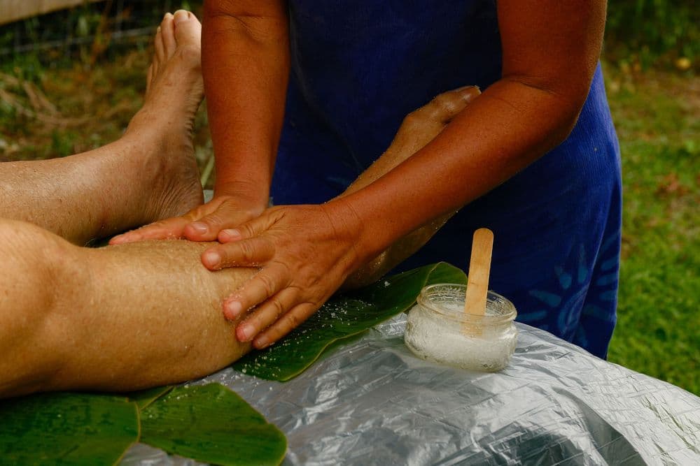 A person receives a massage on their leg outdoors, with hands applying lotion and a jar of cream nearby.