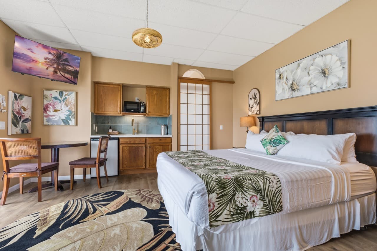 A cozy hotel room featuring a queen bed, kitchenette, and dining area with tropical decor.