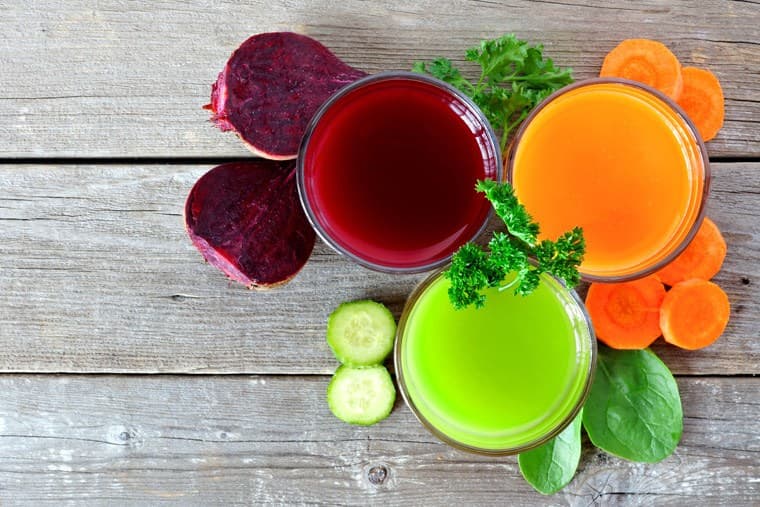 Three colorful fruit and vegetable juices in glass jars, surrounded by fresh ingredients on a wooden surface.