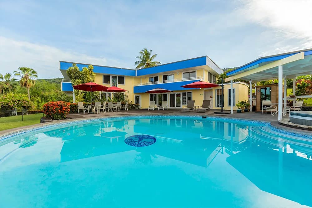 A sunny outdoor pool with a blue-topped house and red umbrellas surrounded by lush greenery.