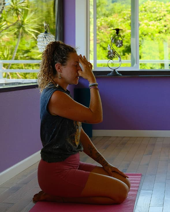 A woman practices a seated breathing exercise on a yoga mat in a brightly lit room.