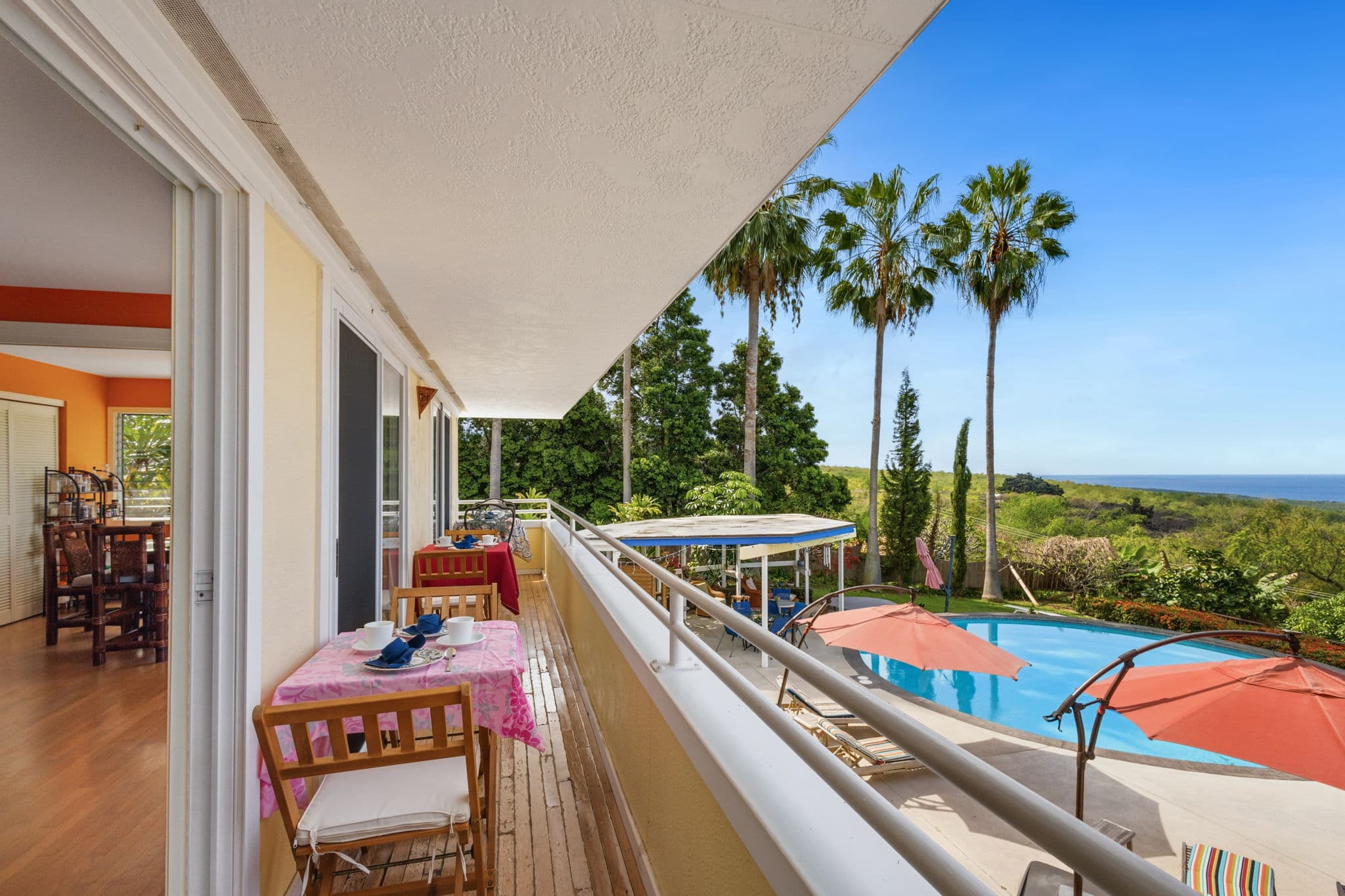 A balcony with a table set for breakfast overlooks a pool and palm trees against a clear blue sky.