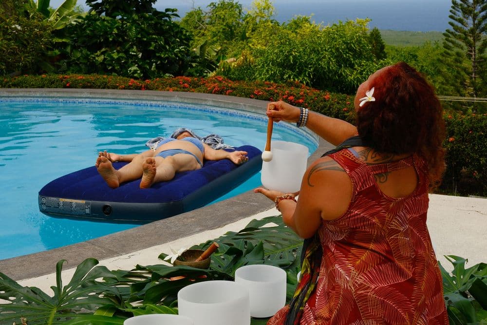A person lounges on a float in a swimming pool while another individual plays crystal bowls nearby.