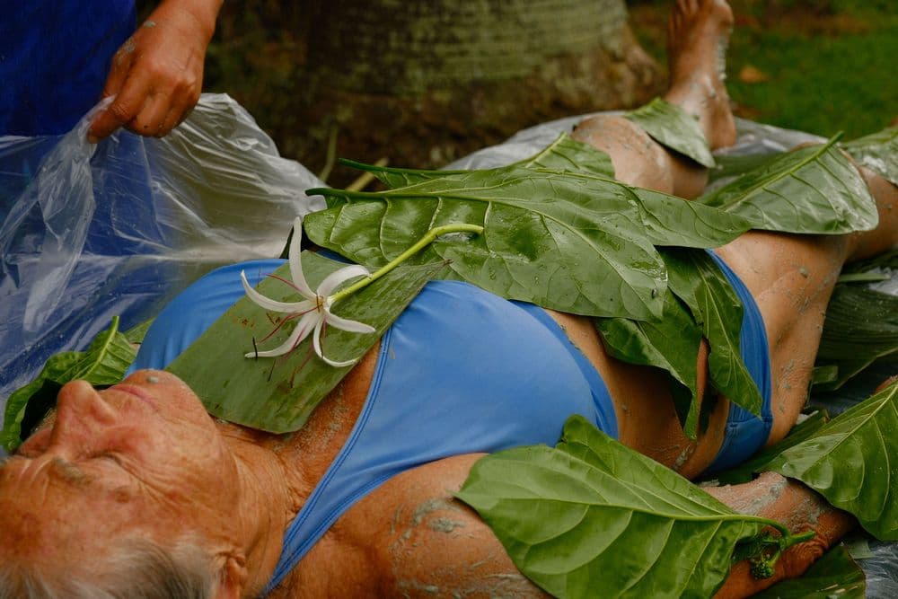 An elderly woman lies on a mat covered with leaves and a flower, while someone applies a substance to her body.