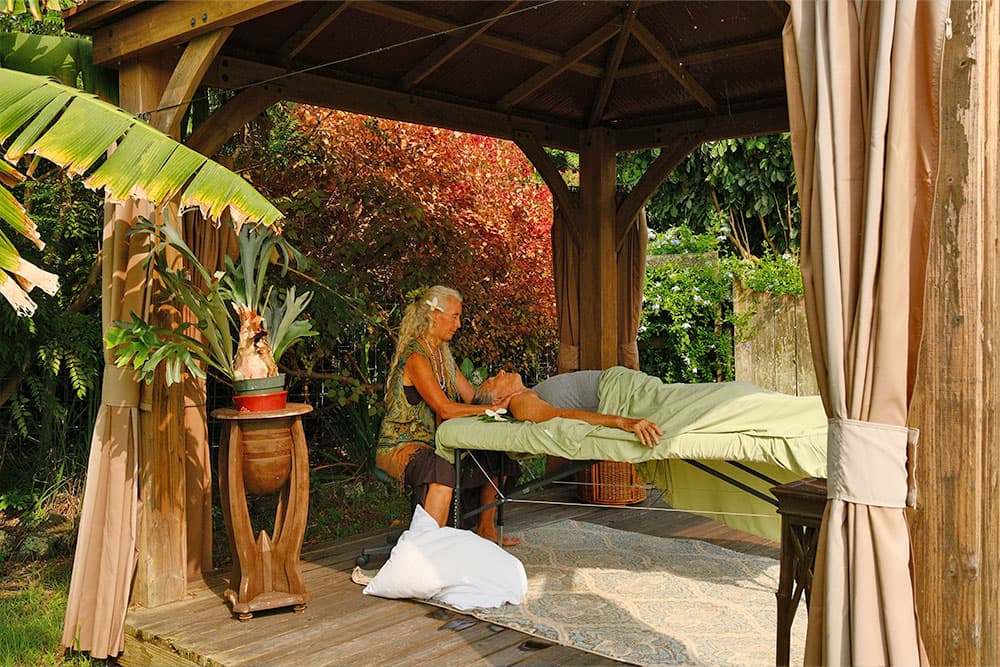 A person gives a massage on a table in a sheltered outdoor gazebo surrounded by lush greenery.
