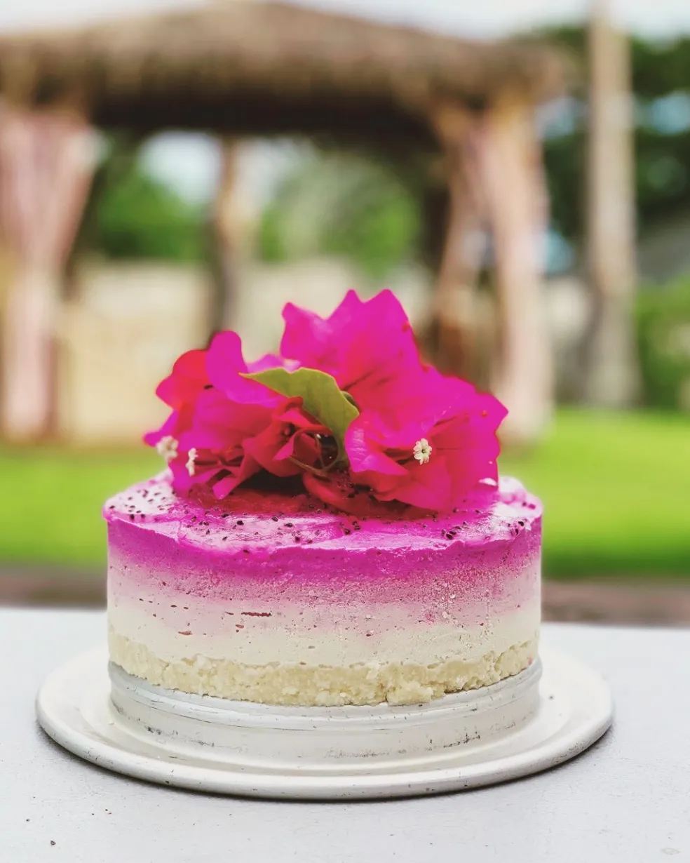 A layered cake topped with vibrant pink flowers, set against a blurred outdoor backdrop.