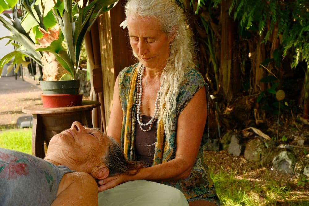 A woman gives a massage to another woman in a serene outdoor setting surrounded by plants.