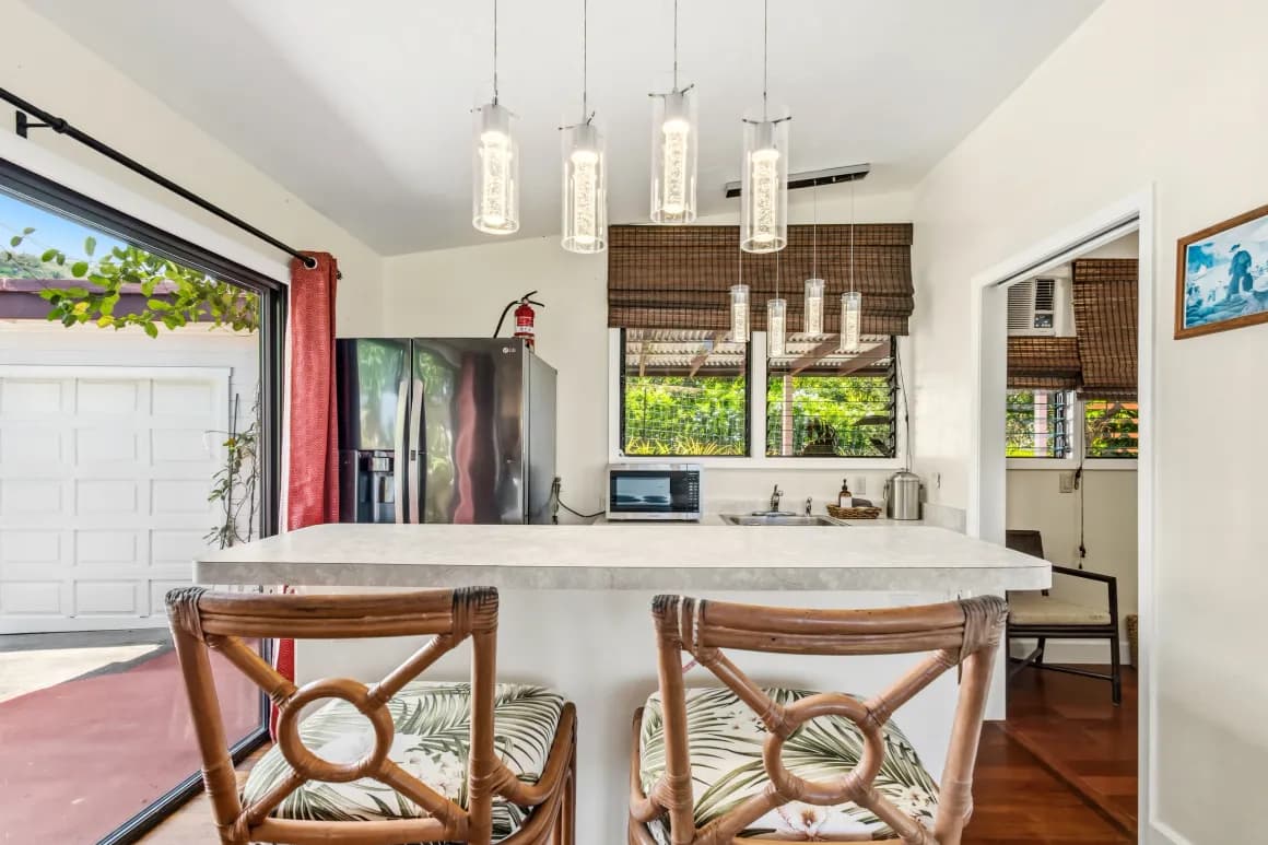 A modern kitchen featuring a marble countertop, woven bar stools, pendant lights, and appliances with a view of the outdoors.