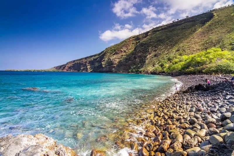 A scenic coastline featuring clear turquoise water and rocky shores against a backdrop of lush green hills.