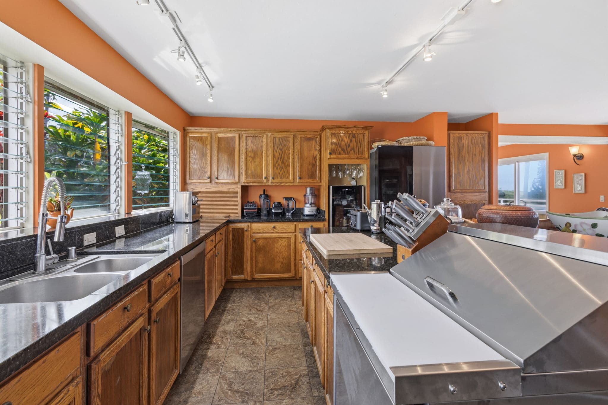 A modern kitchen with wooden cabinets, stainless steel appliances, and large windows revealing greenery outside.