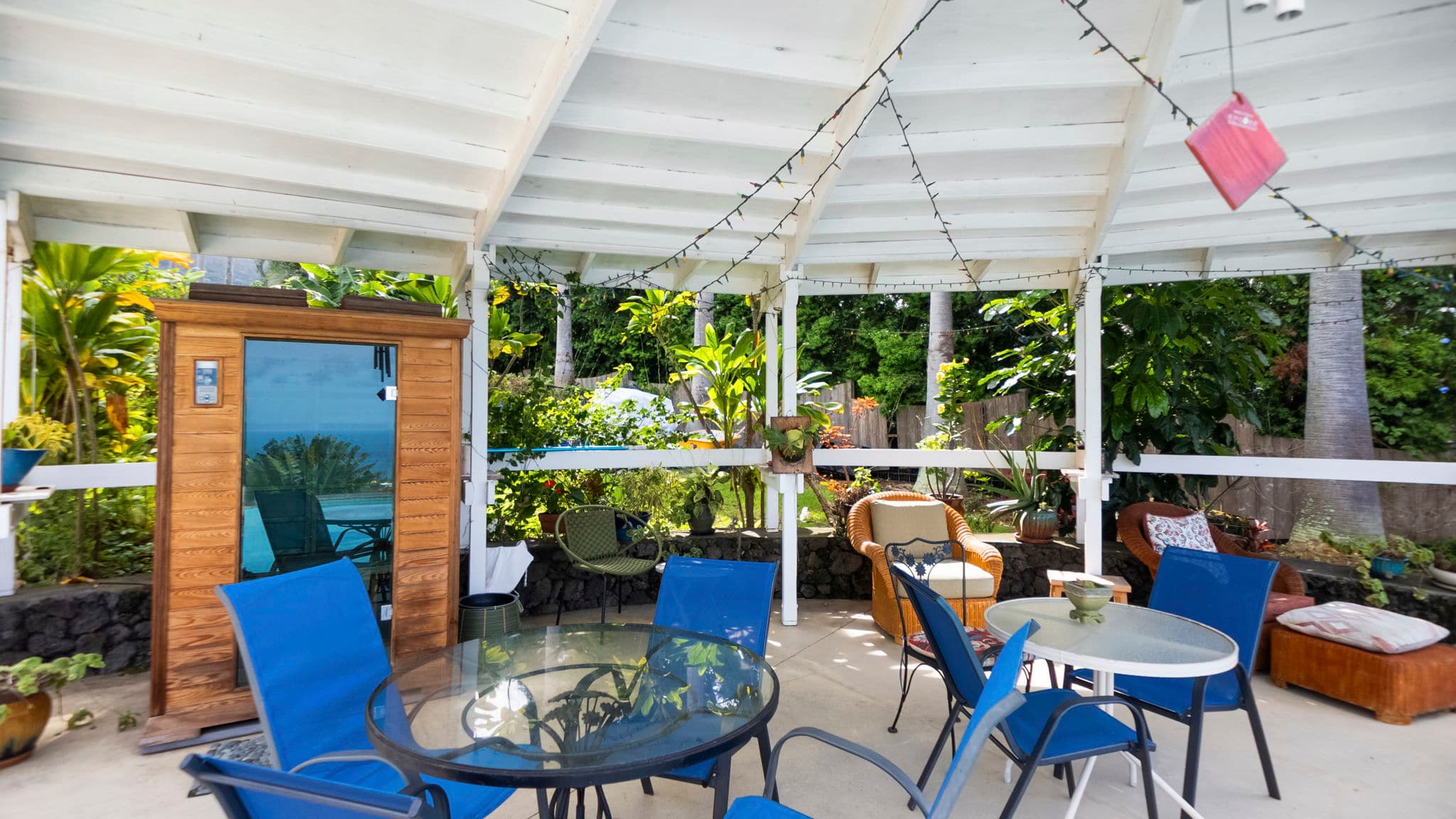 A cozy outdoor seating area with blue chairs, a glass table, and lush greenery under a circular roof.