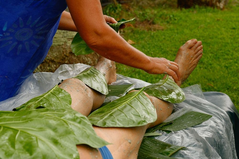A person applies large green leaves to another person's legs while they rest on a plastic sheet outdoors.