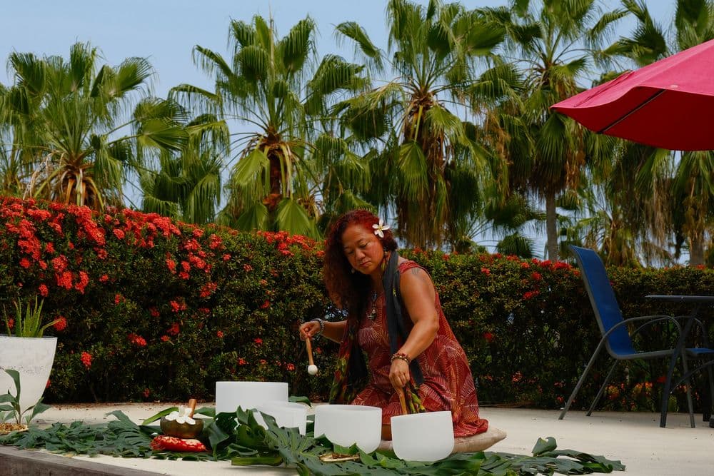 A woman prepares a sound healing ceremony with crystal bowls surrounded by palm trees and vibrant flowers.