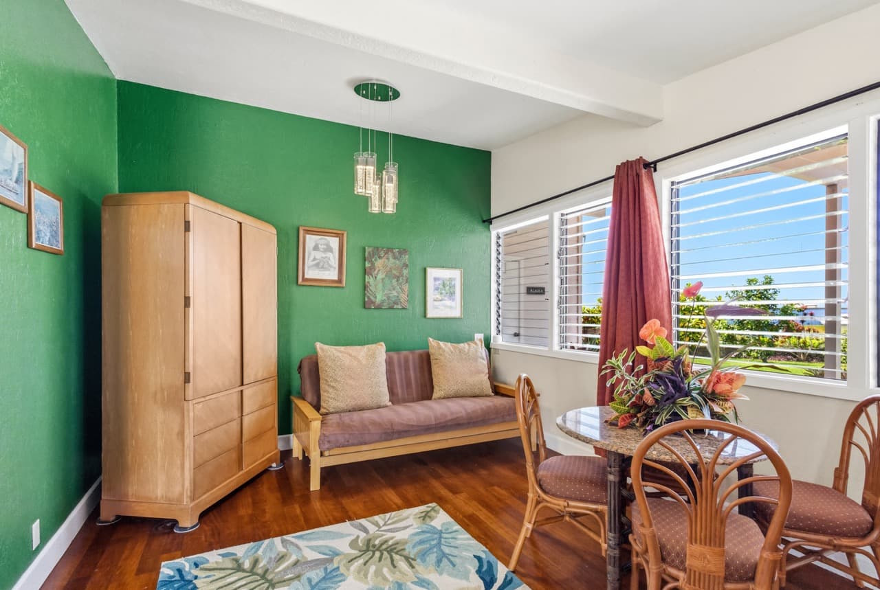 A cozy living space with green walls, a wooden wardrobe, a small dining table, and bright sunlight streaming through the windows.
