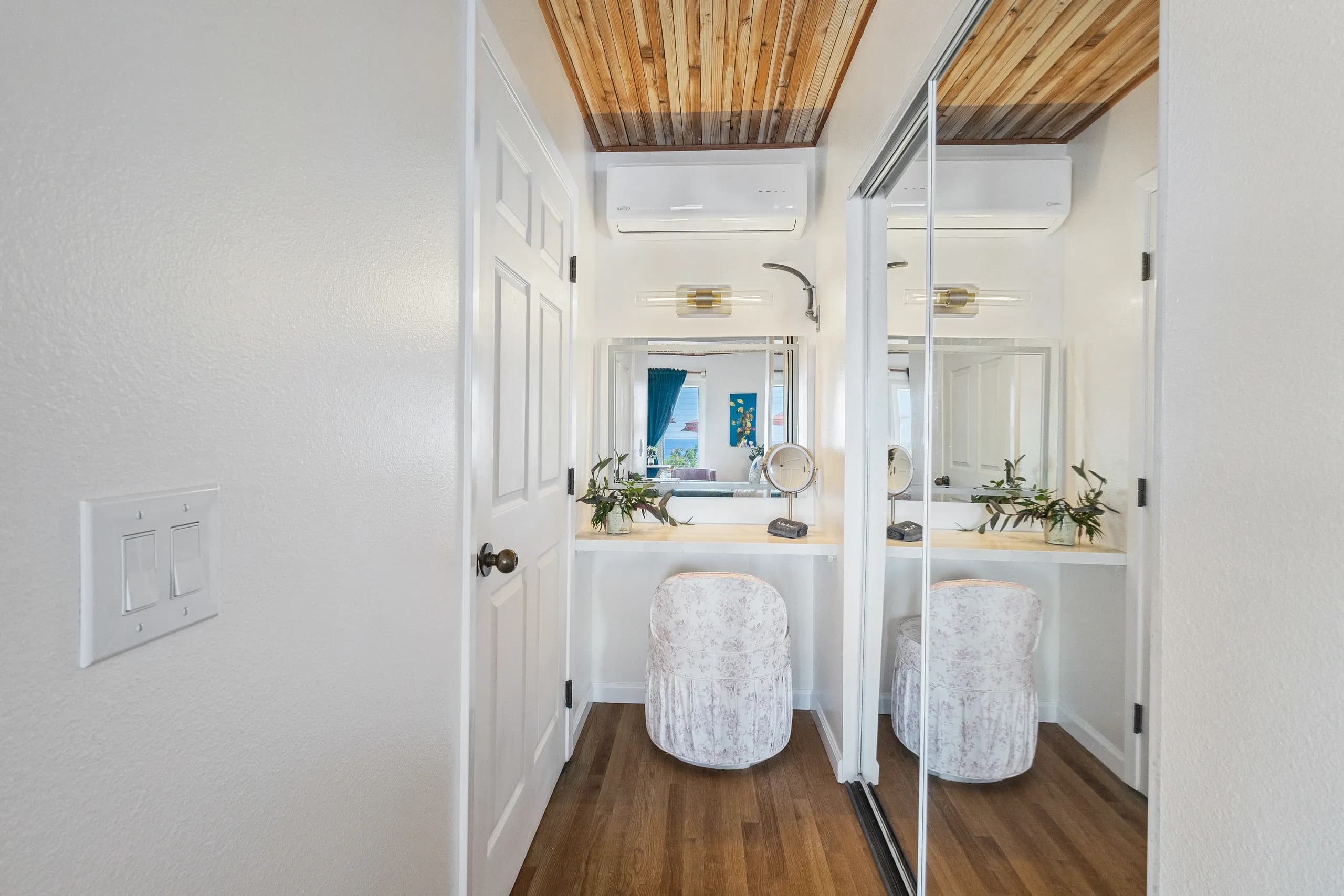 A modern bathroom vanity area with a mirror, stool, and decorative plants, framed by white walls and wooden ceiling.