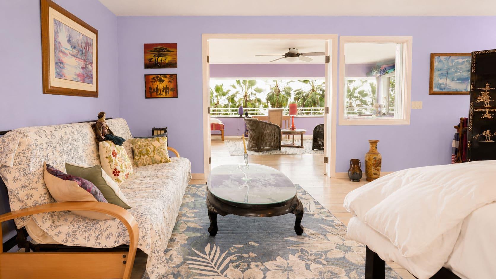 A bright living room with purple walls, a floral rug, a sofa, and a view of outdoor seating through an open door.
