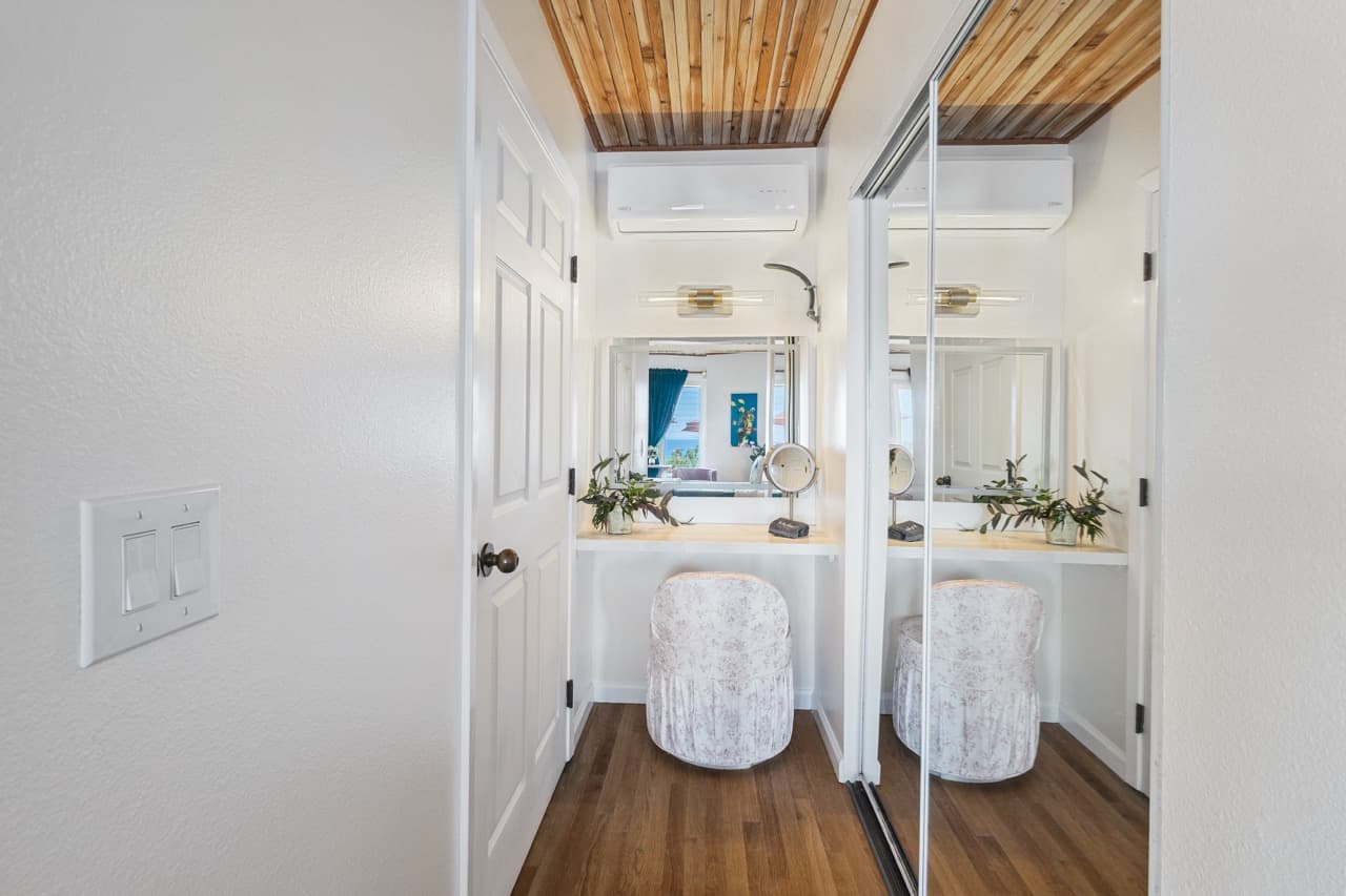 A bright, minimalistic corner vanity area with a mirrored wall, white chair, and wooden ceiling accents.