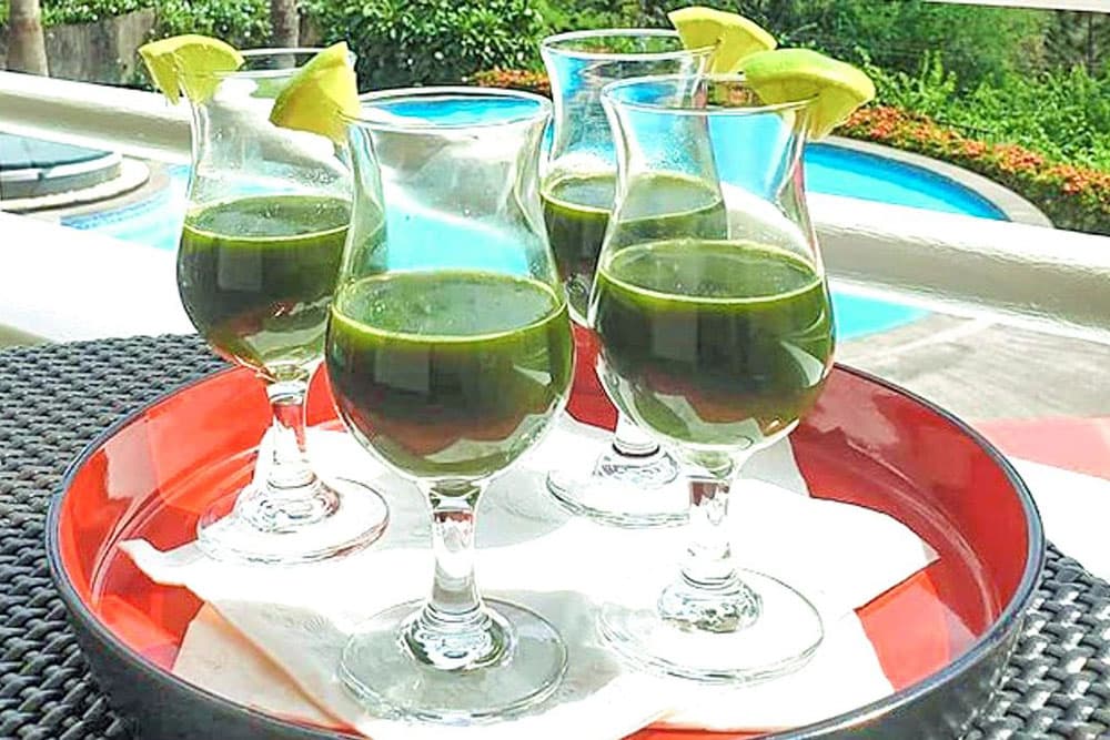 Four glasses of green beverage garnished with lime wedges, served on a red tray beside a pool.