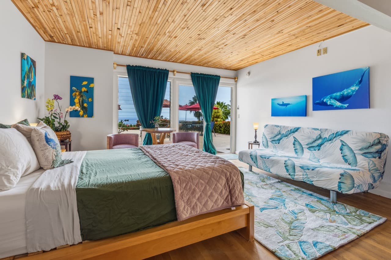A cozy bedroom with a wooden ceiling, green and beige bedding, and artwork, featuring large windows that open to a garden view.