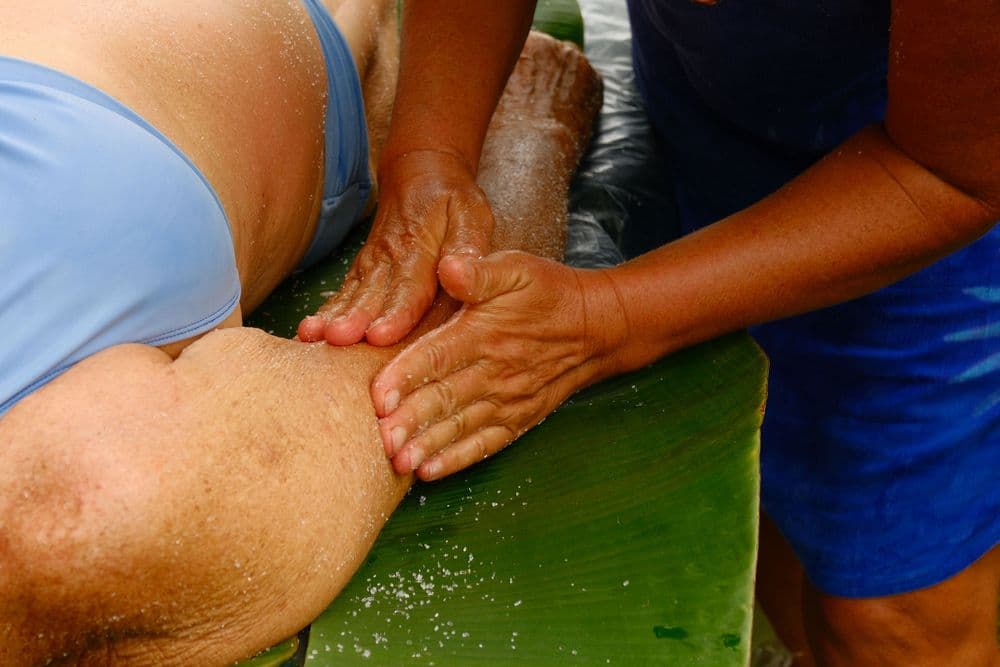A person receives a massage with hands applying pressure to their arm, positioned on a green leaf surface.