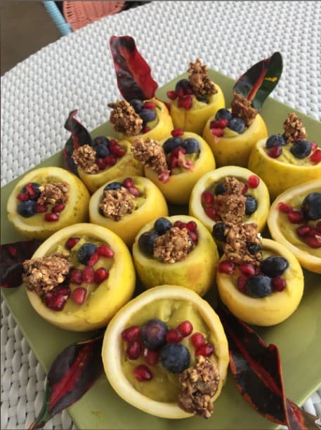 A plate of halved yellow fruits filled with creamy toppings, pomegranate seeds, and blueberries, garnished with crunchy granola.