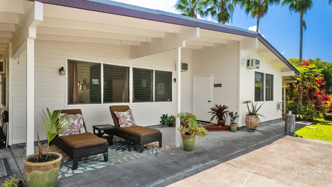 A well-lit, modern porch features two lounge chairs and potted plants alongside a white house.