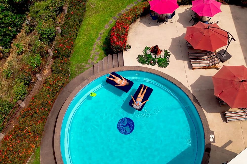 Aerial view of two people lounging in a circular pool surrounded by vibrant greenery and pink umbrellas.