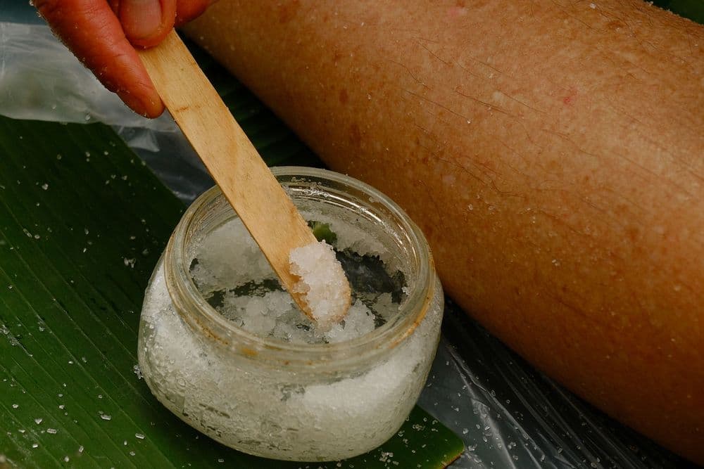A hand with a wooden stick is mixing salt in a jar next to a leg resting on a green leaf.