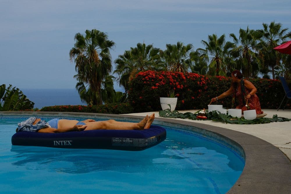 A person relaxes on an inflatable mattress in a pool while another woman prepares flowers nearby, surrounded by palm trees.