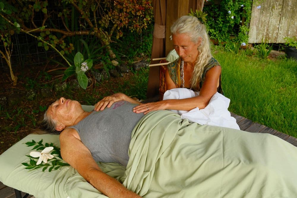 A woman provides a healing touch to a man lying on a massage table surrounded by greenery.