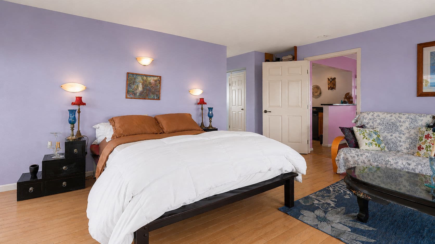 A cozy bedroom with a purple wall, a bed with brown and white bedding, decorative lamps, and a small seating area.