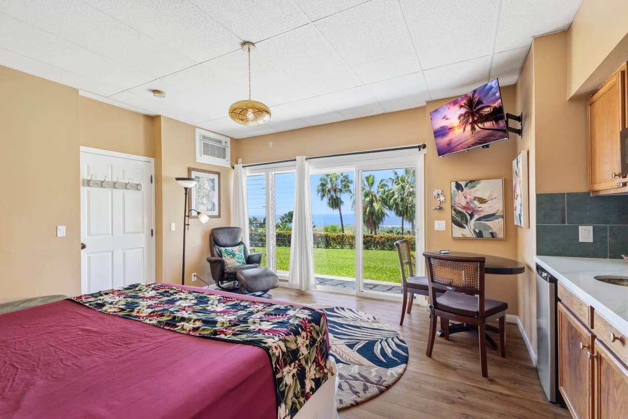 Cozy bedroom with a tropical theme, featuring a bed, armchair, kitchenette, and large windows showcasing a scenic view of palm trees and the ocean.