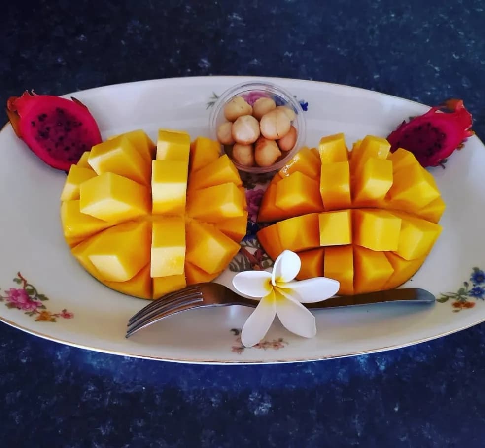 A decorative plate with sliced mangoes, a small bowl of nuts, and dragon fruit, accented by a flower and a fork.