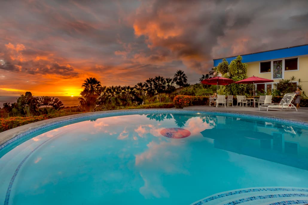 A serene pool reflects a vibrant sunset, surrounded by palm trees and outdoor seating.